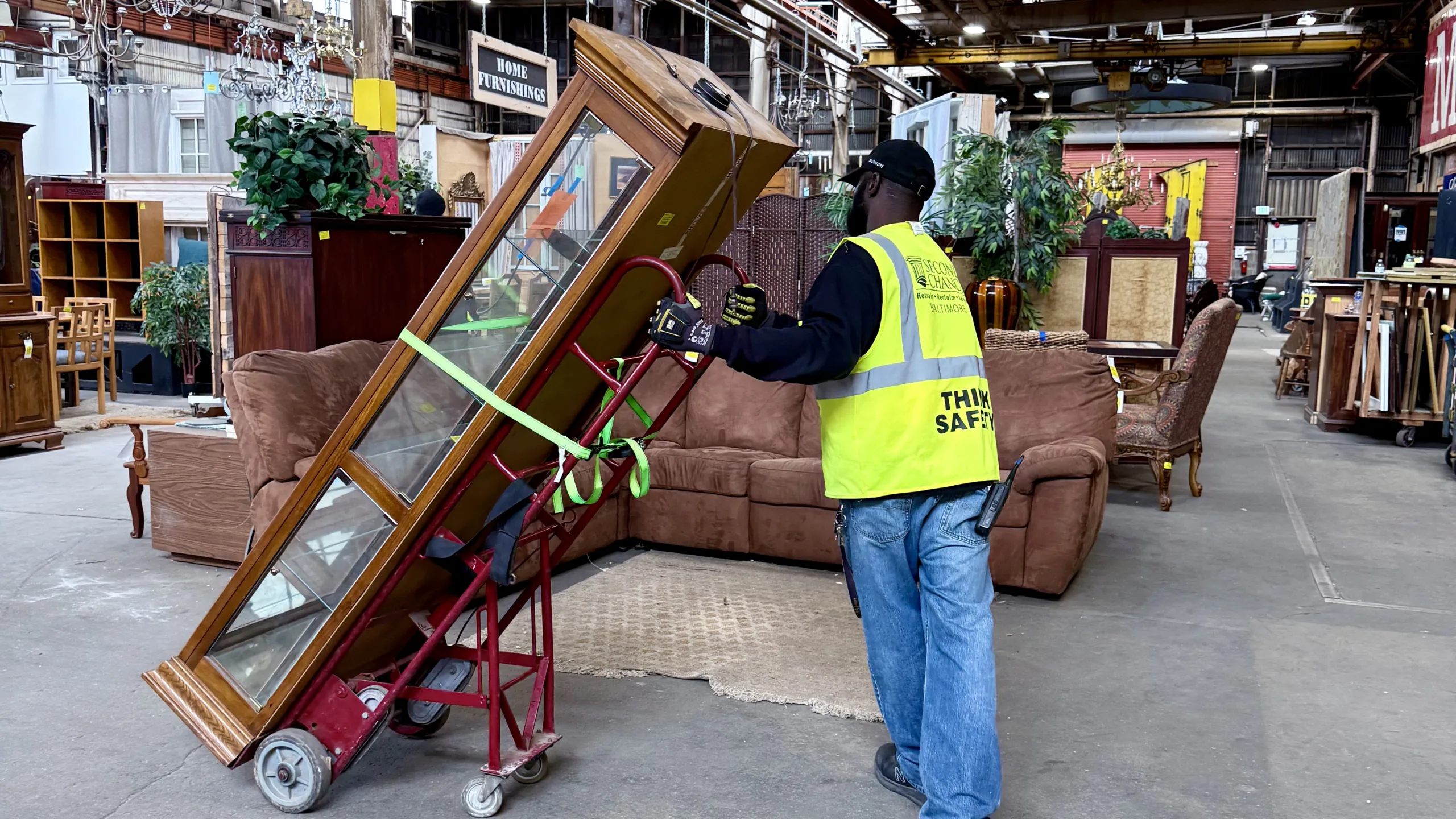 Second Chance Baltimore non-profit employee moving a salvaged vintage furniture display cabinet on a hand truck. The warehouse features a wide array of secondhand home furnishings, unique reclaimed wood items, and other donated goods.