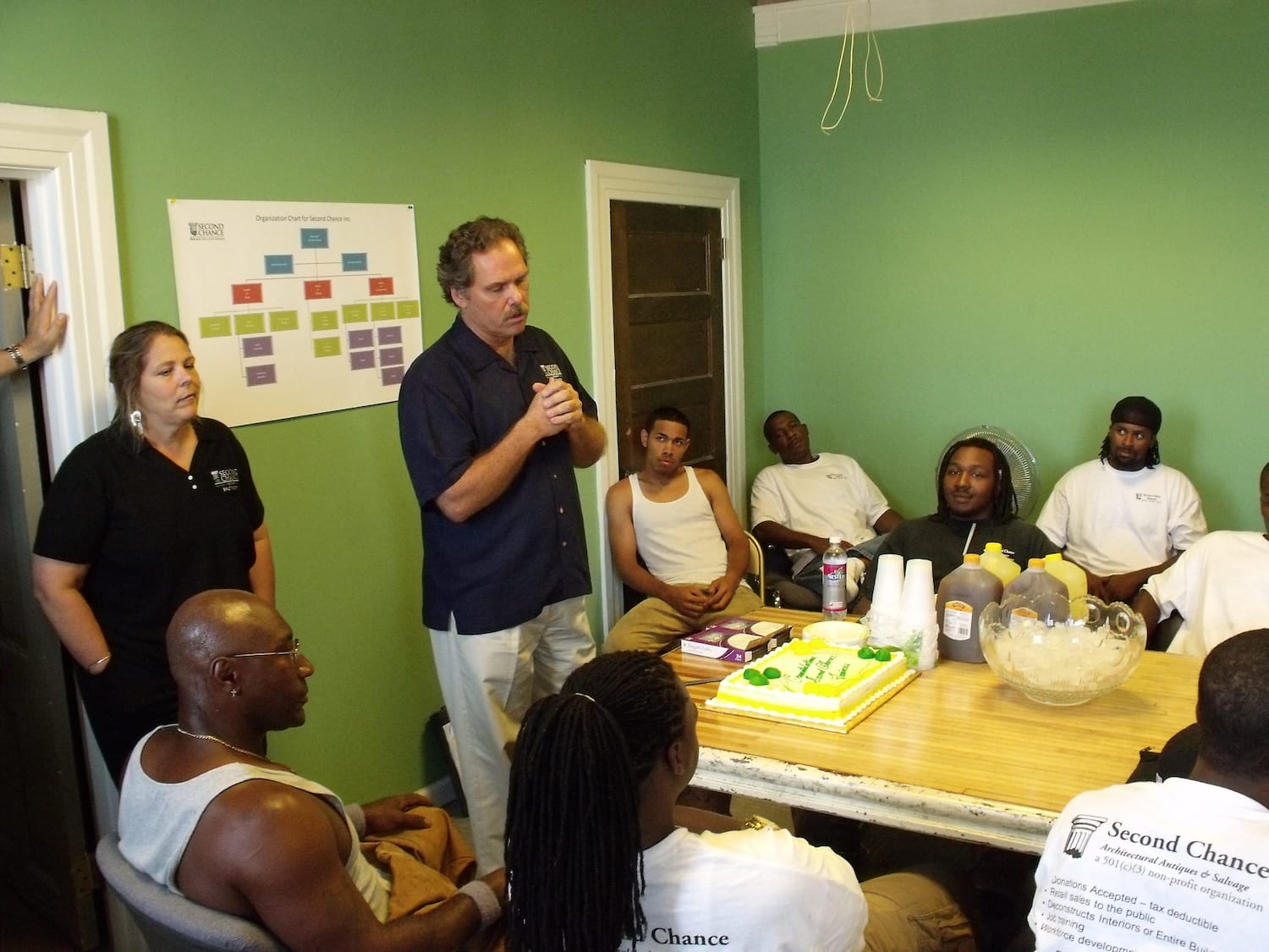 Internal meeting at Second Chance, a Baltimore non-profit, with staff and participants gathered around a wooden table with a celebration cake. The group reviews the organization's mission to salvage and resell used building materials, vintage furniture, and other secondhand goods. An organizational chart for the Baltimore non-profit is displayed on the green wall.