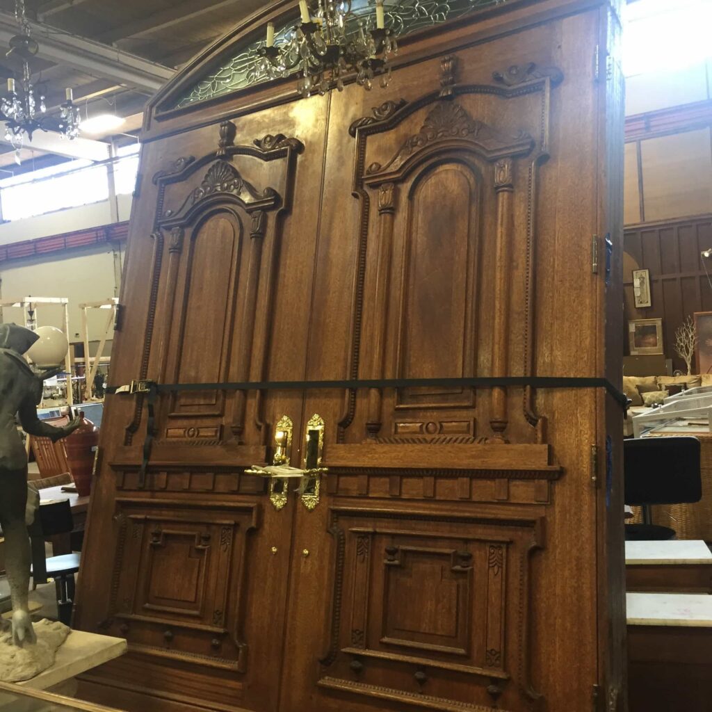 Ornate salvaged wooden double doors featuring intricate carvings, a textured glass transom, and antique gold hardware. Discover unique secondhand building materials and vintage finds at Second Chance, a Baltimore non-profit.