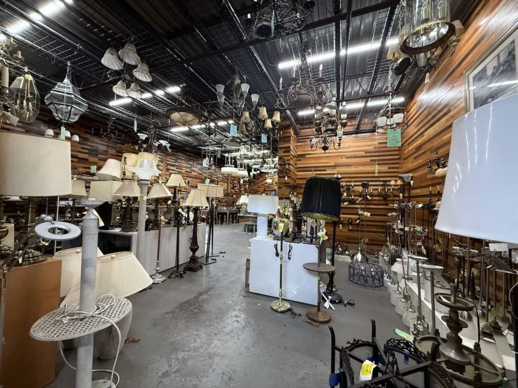 Extensive collection of salvaged and secondhand lighting, including vintage chandeliers, floor lamps, and table lamps, displayed at a Baltimore non-profit thrift store. Features a wall of reclaimed wood paneling, offering unique home decor and used building materials.