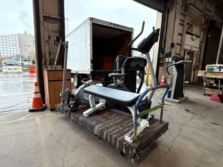 An outbound donation of secondhand exercise equipment, including an elliptical, rowing machine, and weight bench, alongside a used wooden cabinet, getting ready to be loaded onto a truck for Living Classrooms.