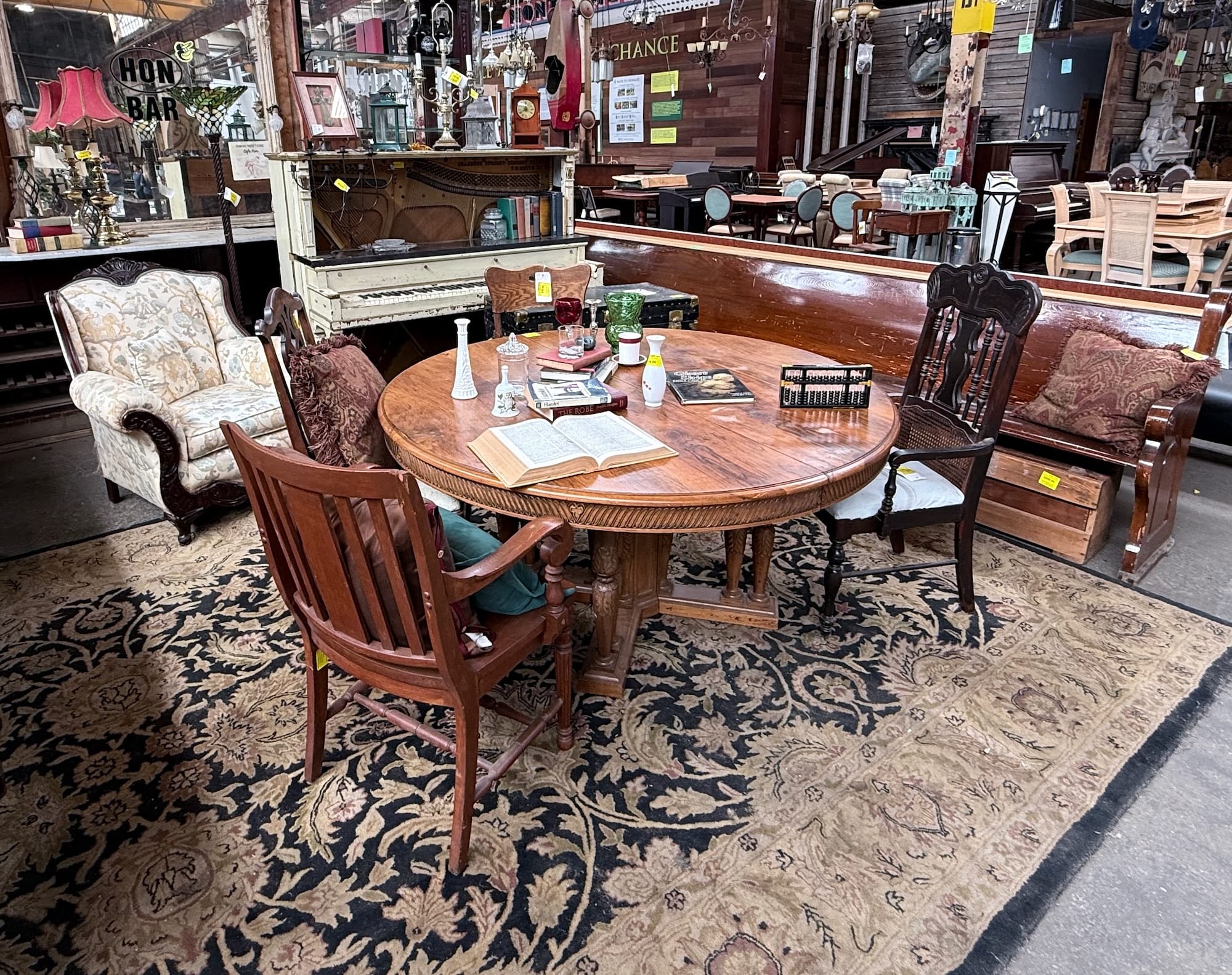 Interior of Second Chance, Inc., a Baltimore non-profit, featuring a round reclaimed wood dining table with vintage chairs on an ornate secondhand rug. Surrounding unique finds include an upholstered armchair and a distressed white piano, showcasing sustainable home decor and secondhand furniture options.