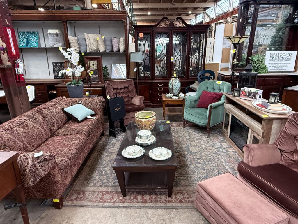 Interior view of Second Chance, Inc., a Baltimore non-profit, showcasing a diverse collection of secondhand and vintage furniture. Featured pieces include a patterned sofa, two upholstered armchairs, a dark wood display cabinet, and a coffee table with decorative items. This unique assortment of home decor, potentially including salvaged or reclaimed wood pieces, offers sustainable options for any home improvement project.